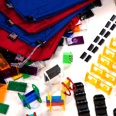 Atwood Toys Bulk Dominoes Super Mega Kit Made in USA canvas storage bags in red and blue with clear and colored dominoes, domino keychain, and accessories scattered around